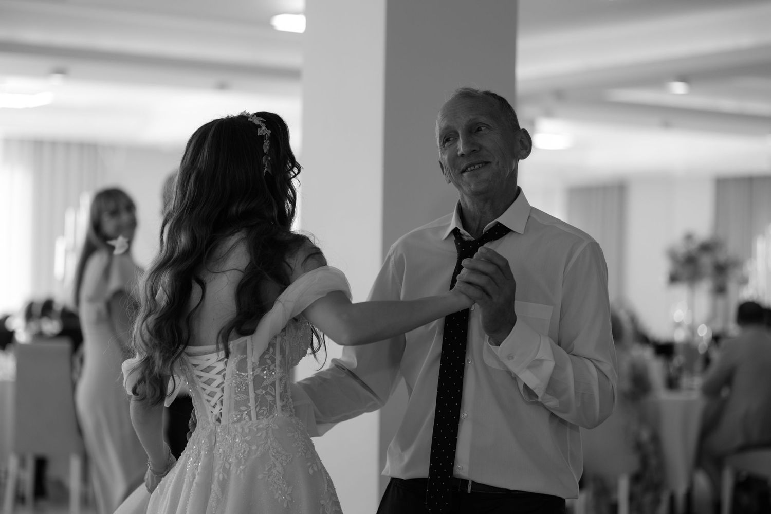 A black and white photo shows a couple dancing together at a wedding reception in a brightly lit venue.