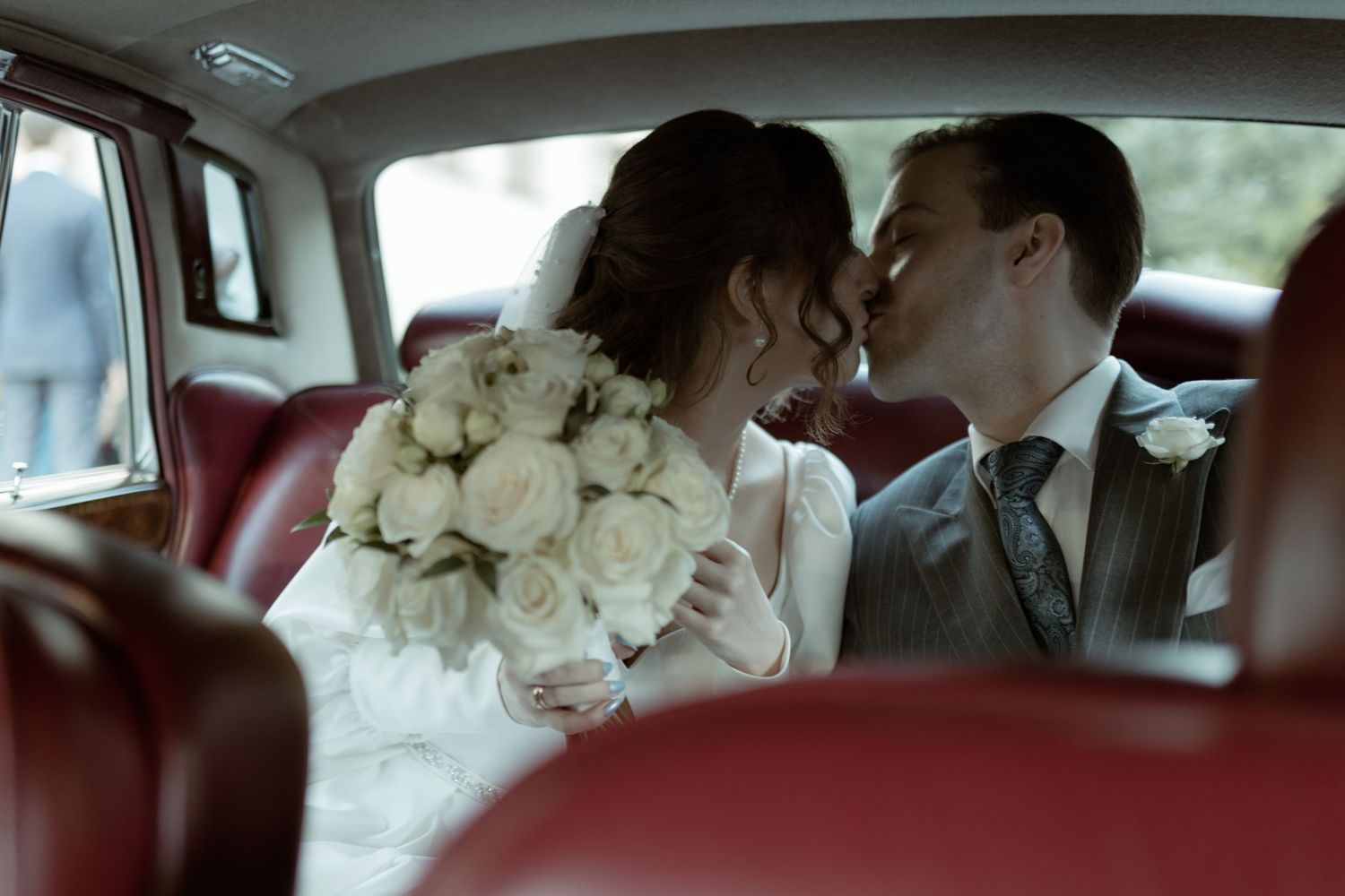 Couple shares intimate moment in red leather backseat of vintage car with white rose bouquet.
