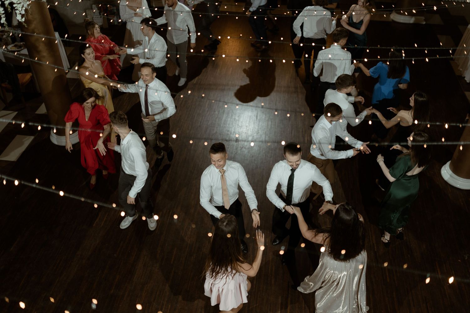 Overhead view of wedding guests dancing on a wooden floor illuminated by string lights at a reception party.