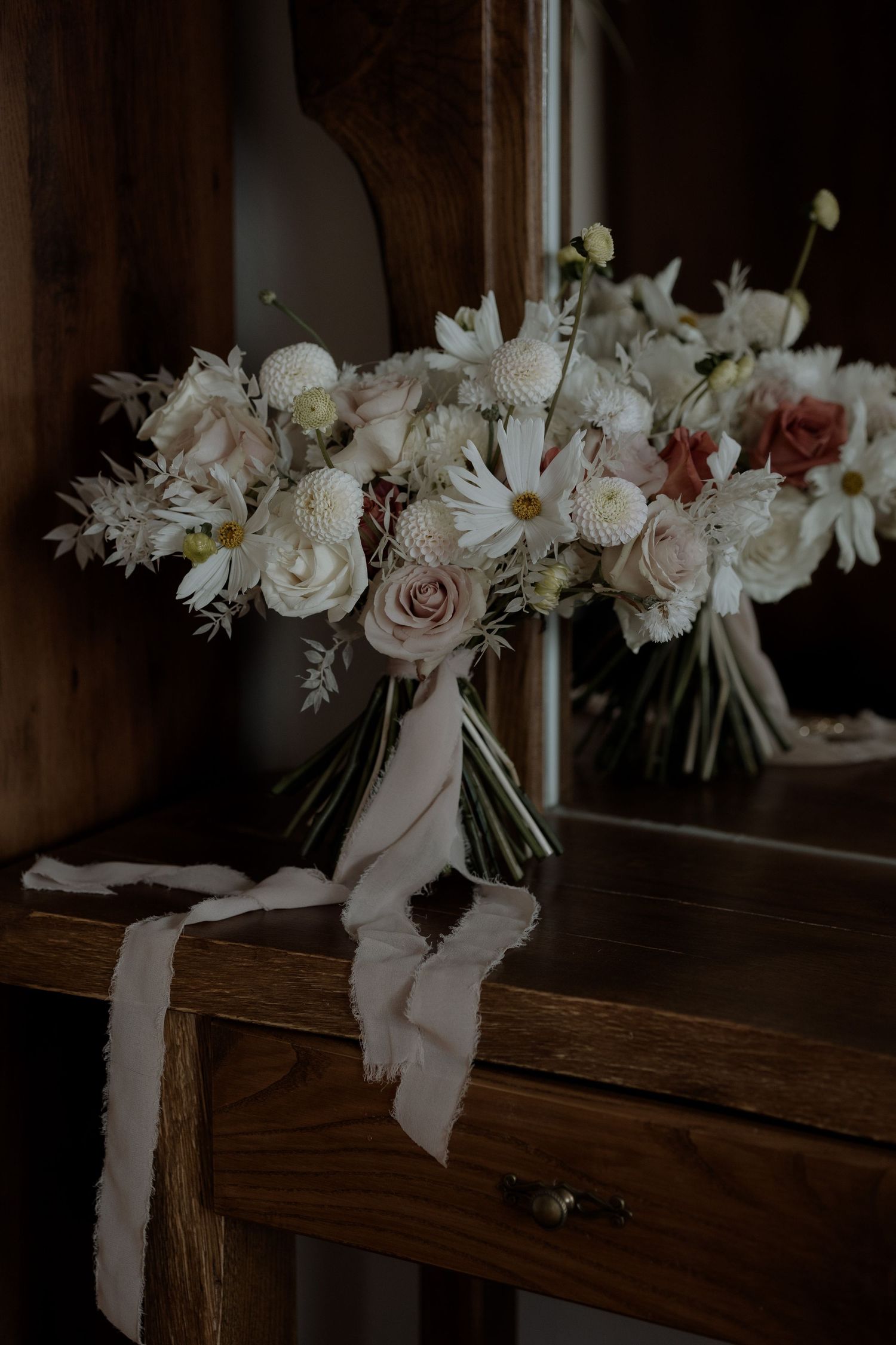A moody wedding bouquet with white and blush roses sits on a dark wooden surface with flowing ribbons.