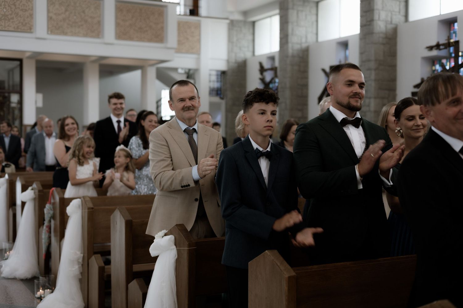 Attendees stand in church pews during a formal ceremony in an elegant interior setting.