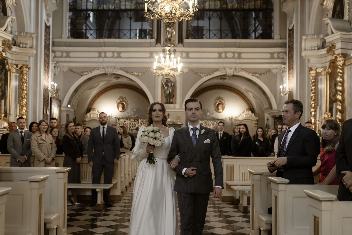 Wedding ceremony taking place in an elegant historic church with ornate architecture and crystal chandeliers.