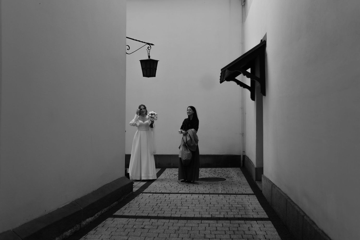 Two people stand in a narrow corridor with white walls and a hanging lantern, creating a dramatic black and white scene.