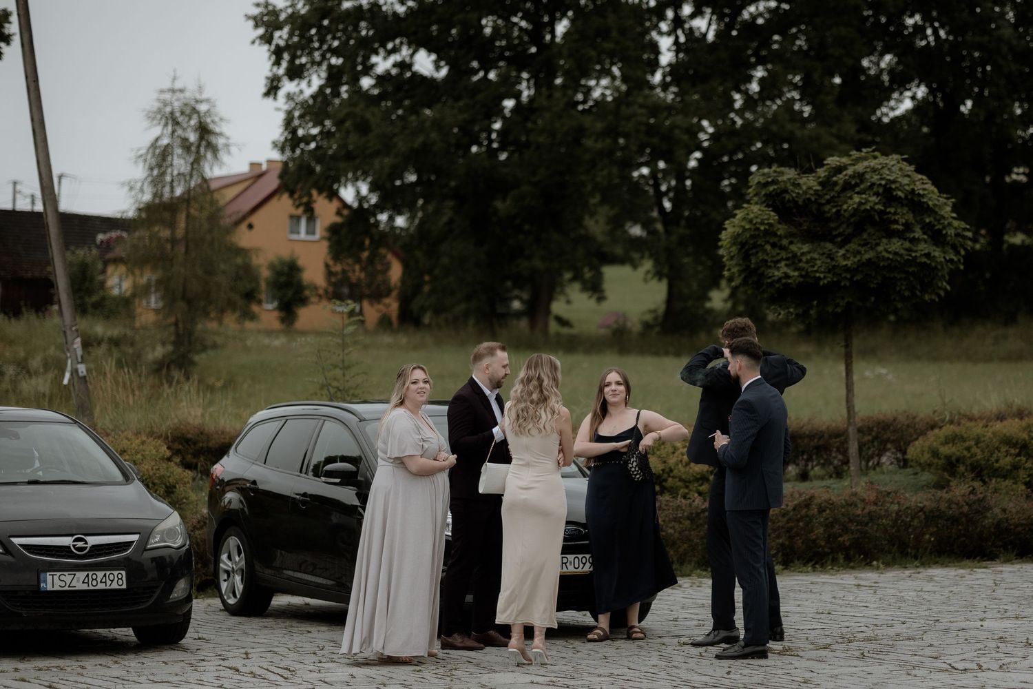 A small group gathers outdoors for a ceremony in front of a parked black car on a cloudy day.