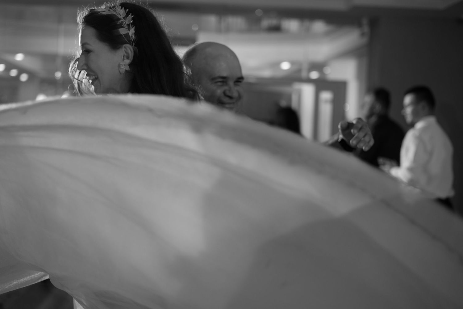 A black and white photo captures people at a wedding celebration with flowing fabric in the foreground.