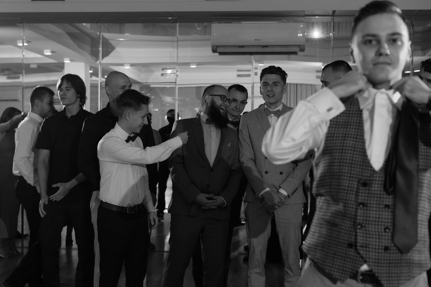 A black and white photograph shows people gathered at an indoor event wearing formal attire including vests and suits.