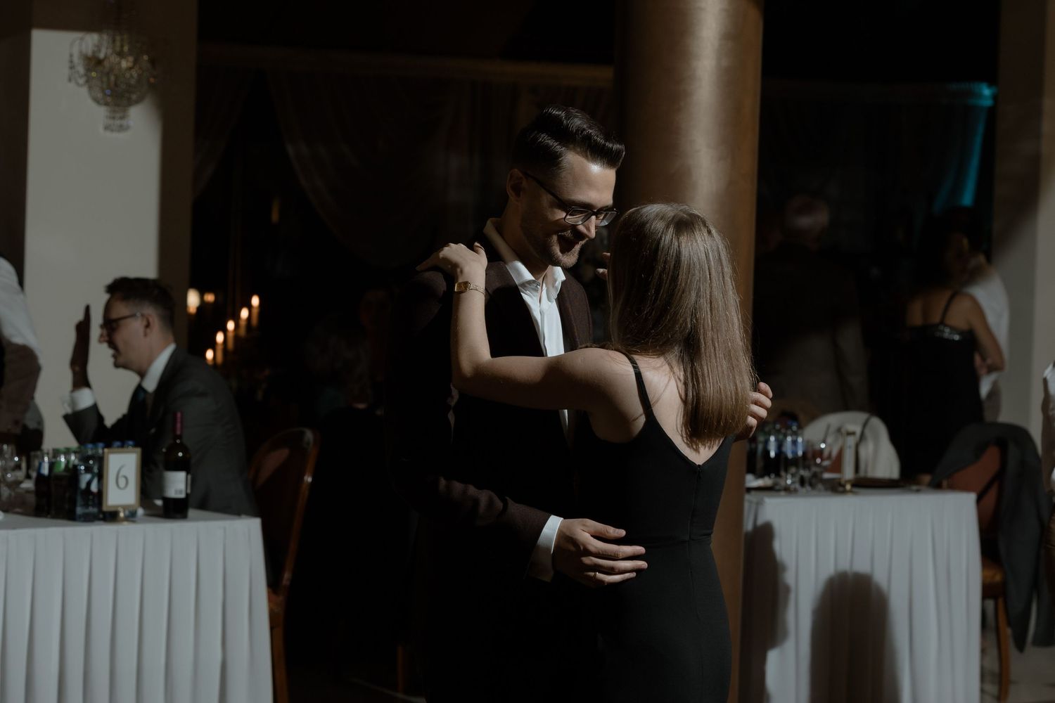 A couple shares an intimate dance at a wedding reception with tables and decor visible in the background.