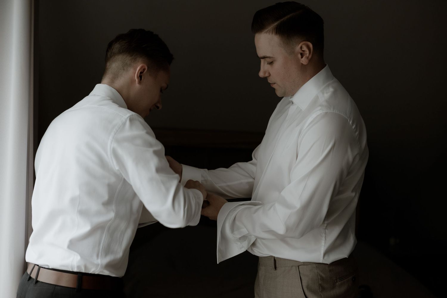 Two people in white dress shirts help each other with formal attire preparations in a dimly lit room.