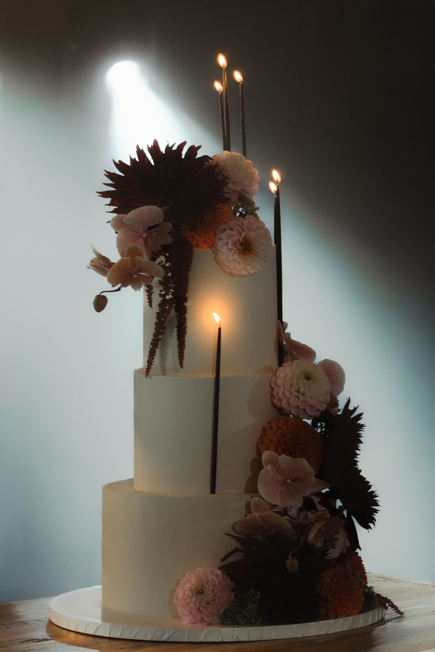 A white tiered wedding cake decorated with burgundy and blush pink flowers captured from different angles in moody lighting.