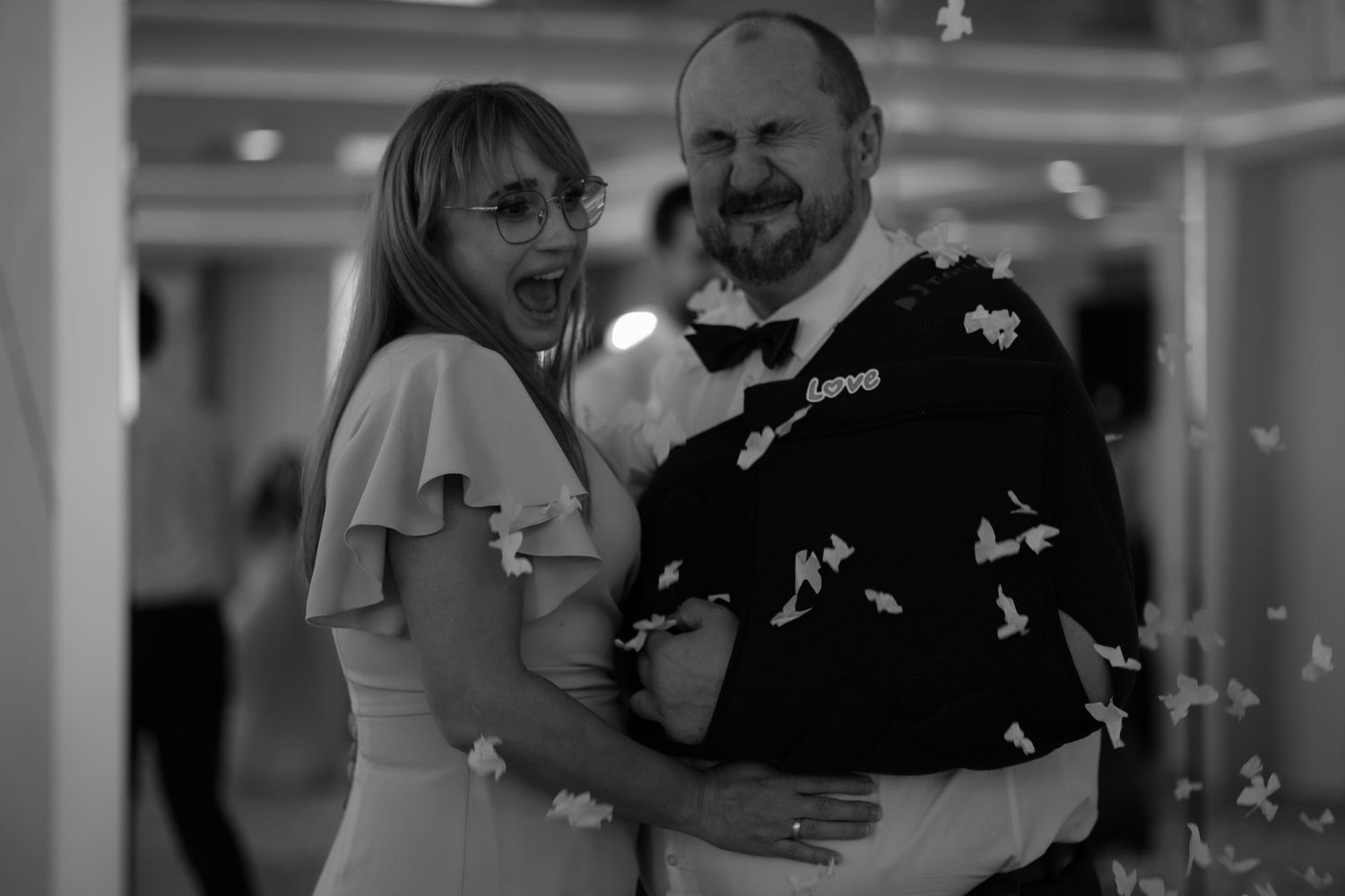 A couple dances together at a wedding reception while confetti falls around them in black and white.