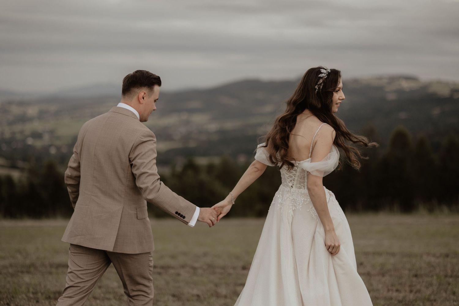 A couple in wedding attire holds hands while walking across a scenic hilltop meadow with mountains in the background.
