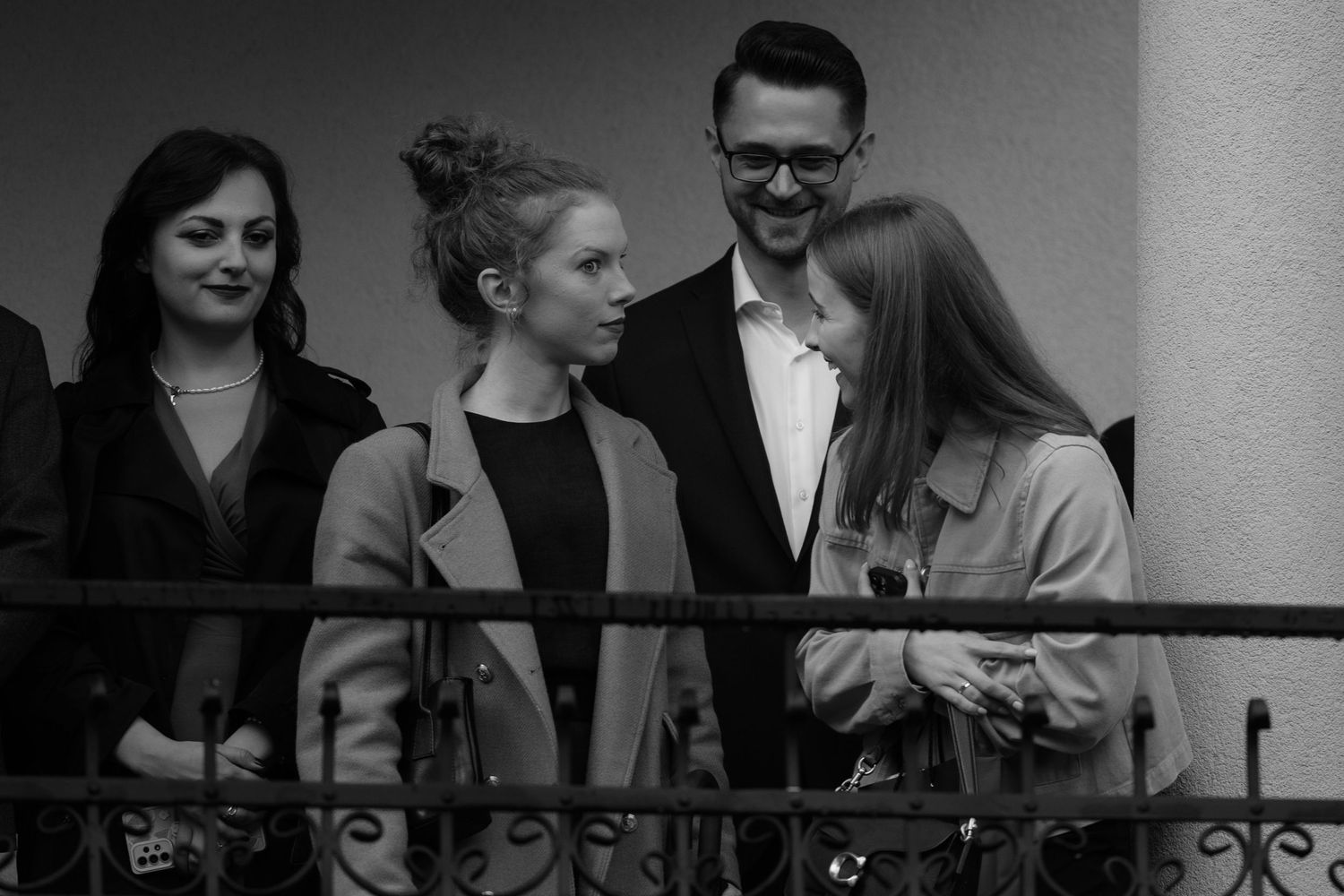 A black and white photograph shows a group of people chatting and smiling while standing on a balcony.