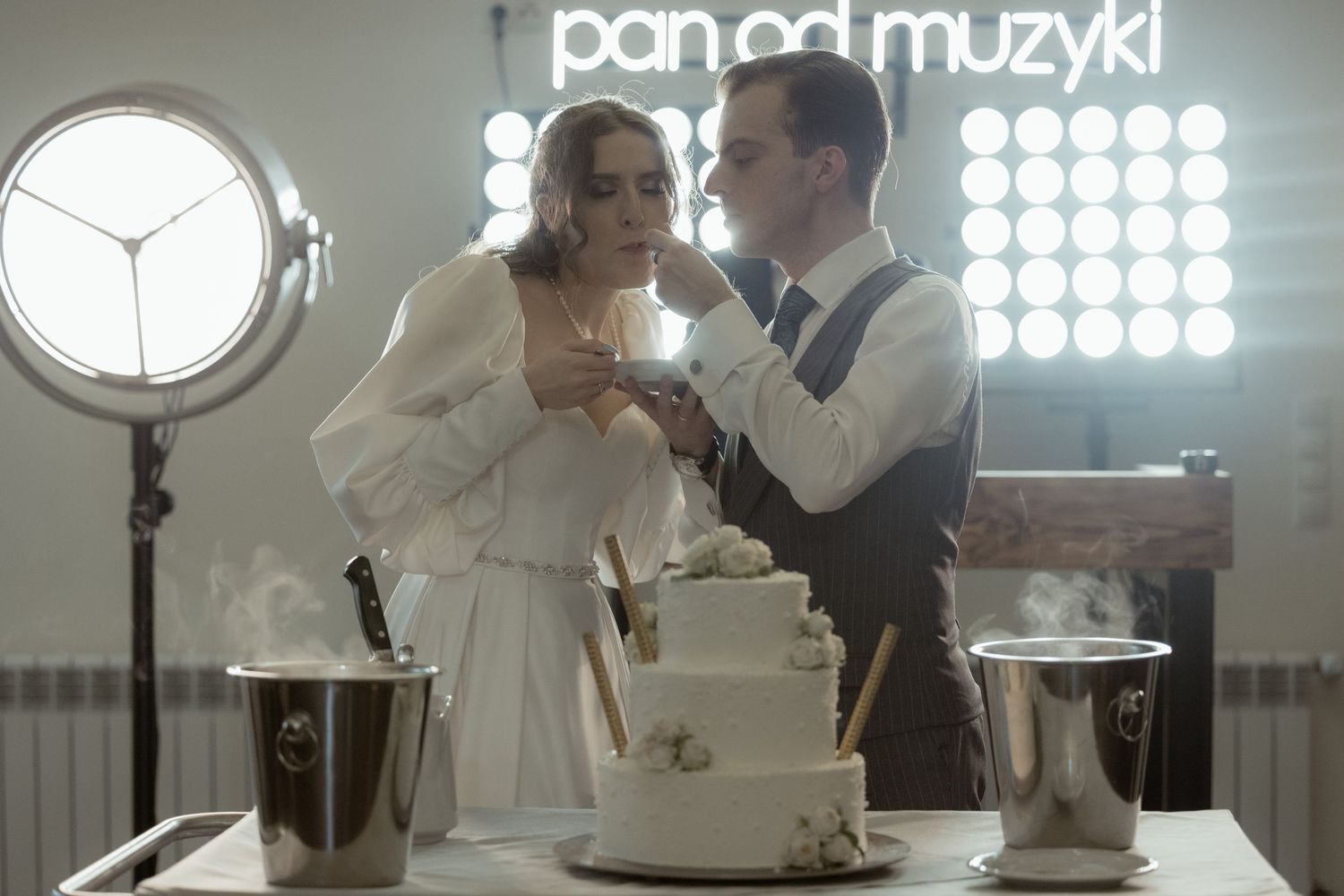 A bride and groom share a romantic moment next to their wedding cake under vintage lighting at their reception.