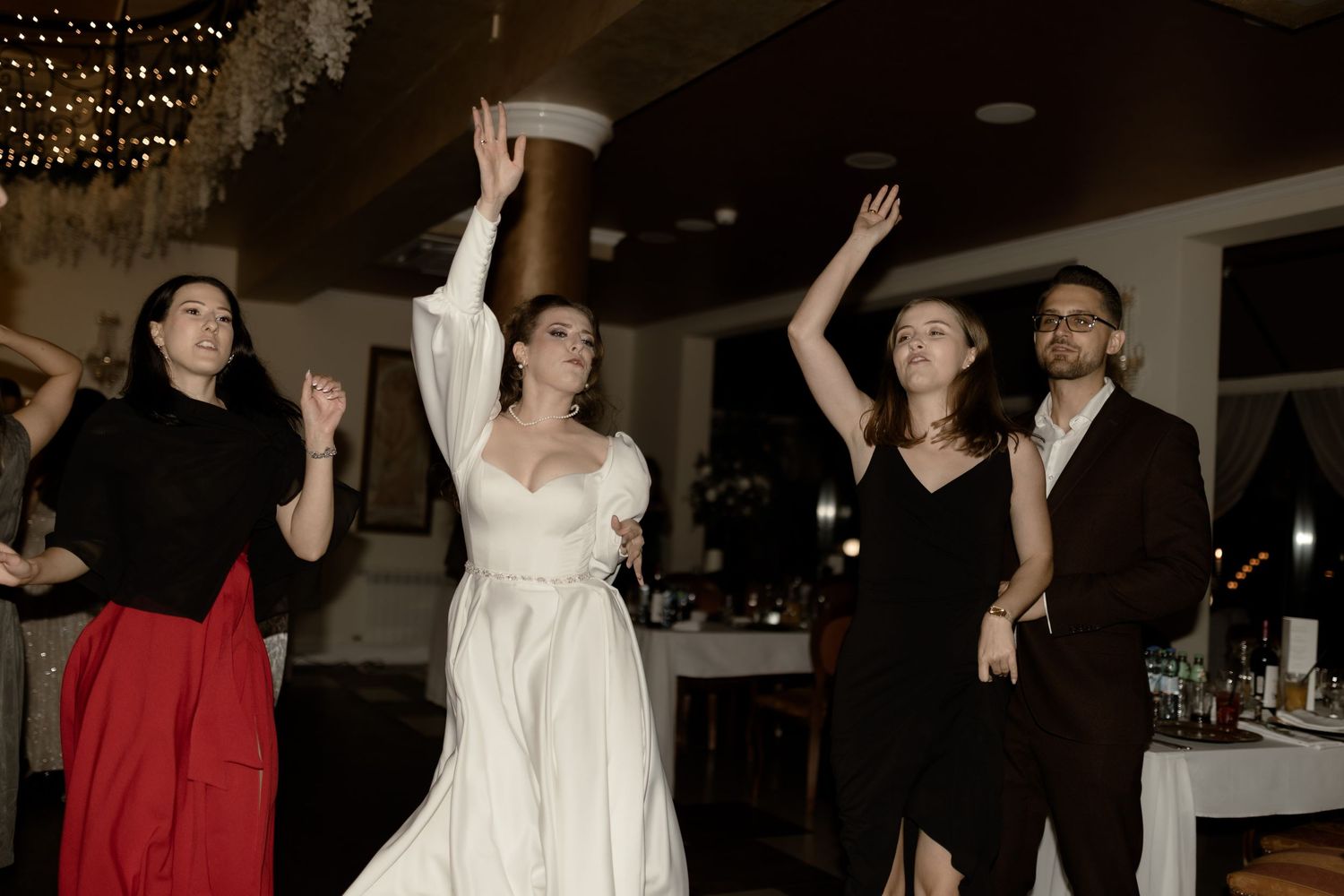 Wedding guests and couple dancing together during a reception celebration with venue lighting in the background.