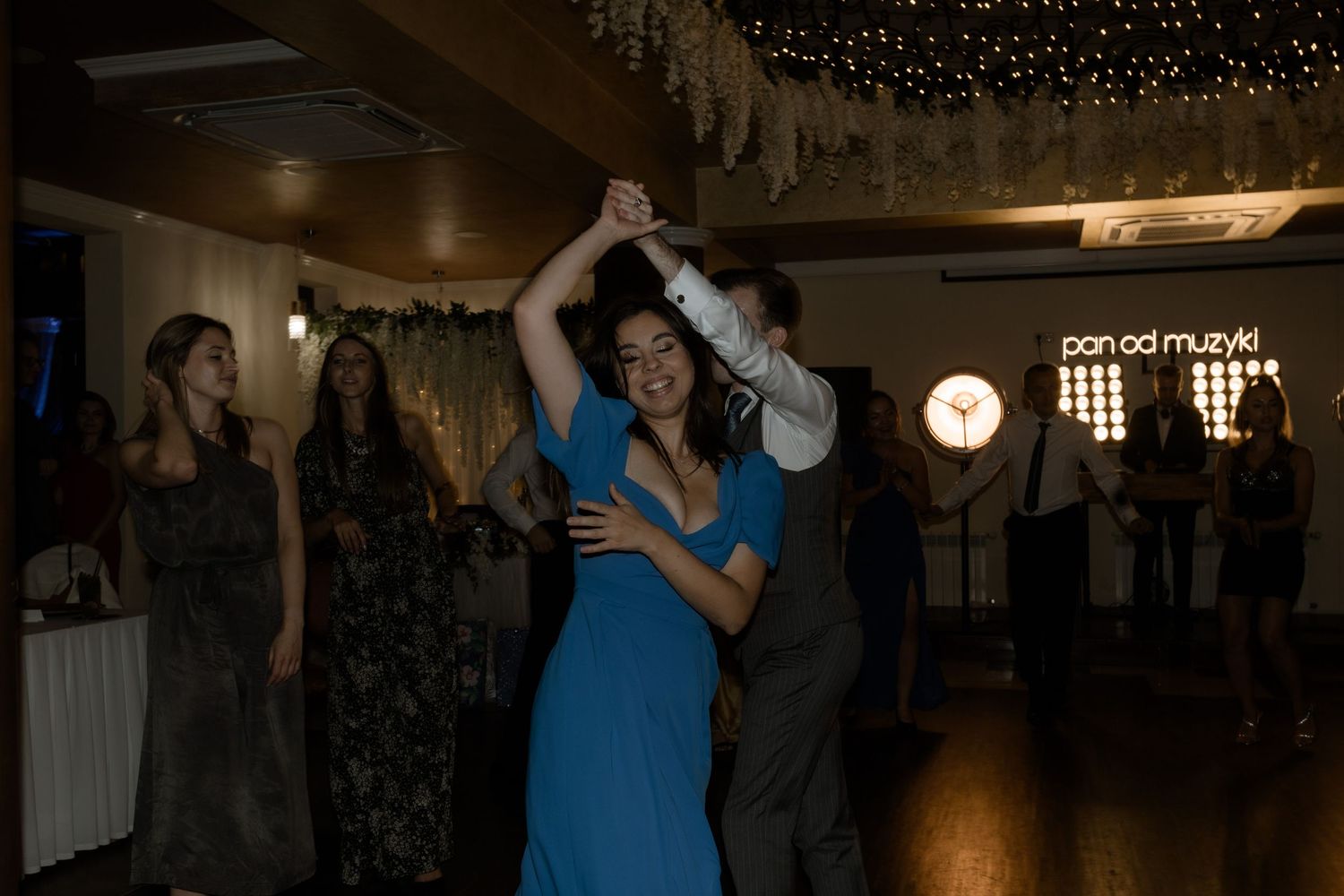 A couple spins on the dance floor at an indoor reception venue illuminated with twinkling lights.