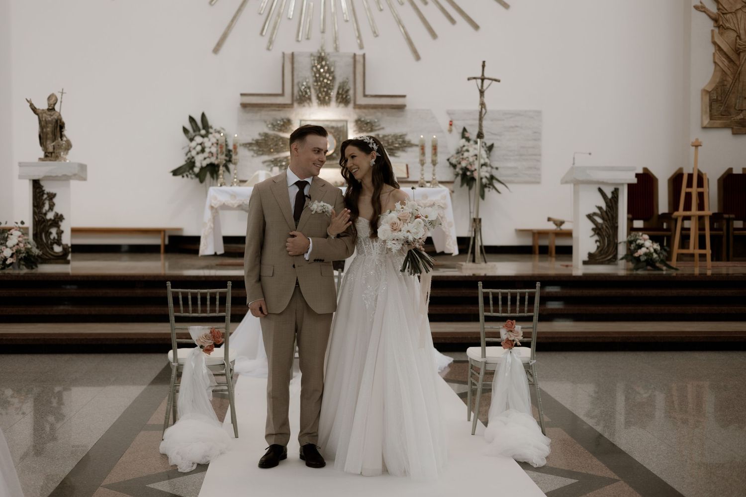 A couple stands at the altar of an elegant church interior with white floral arrangements and decorative crosses.