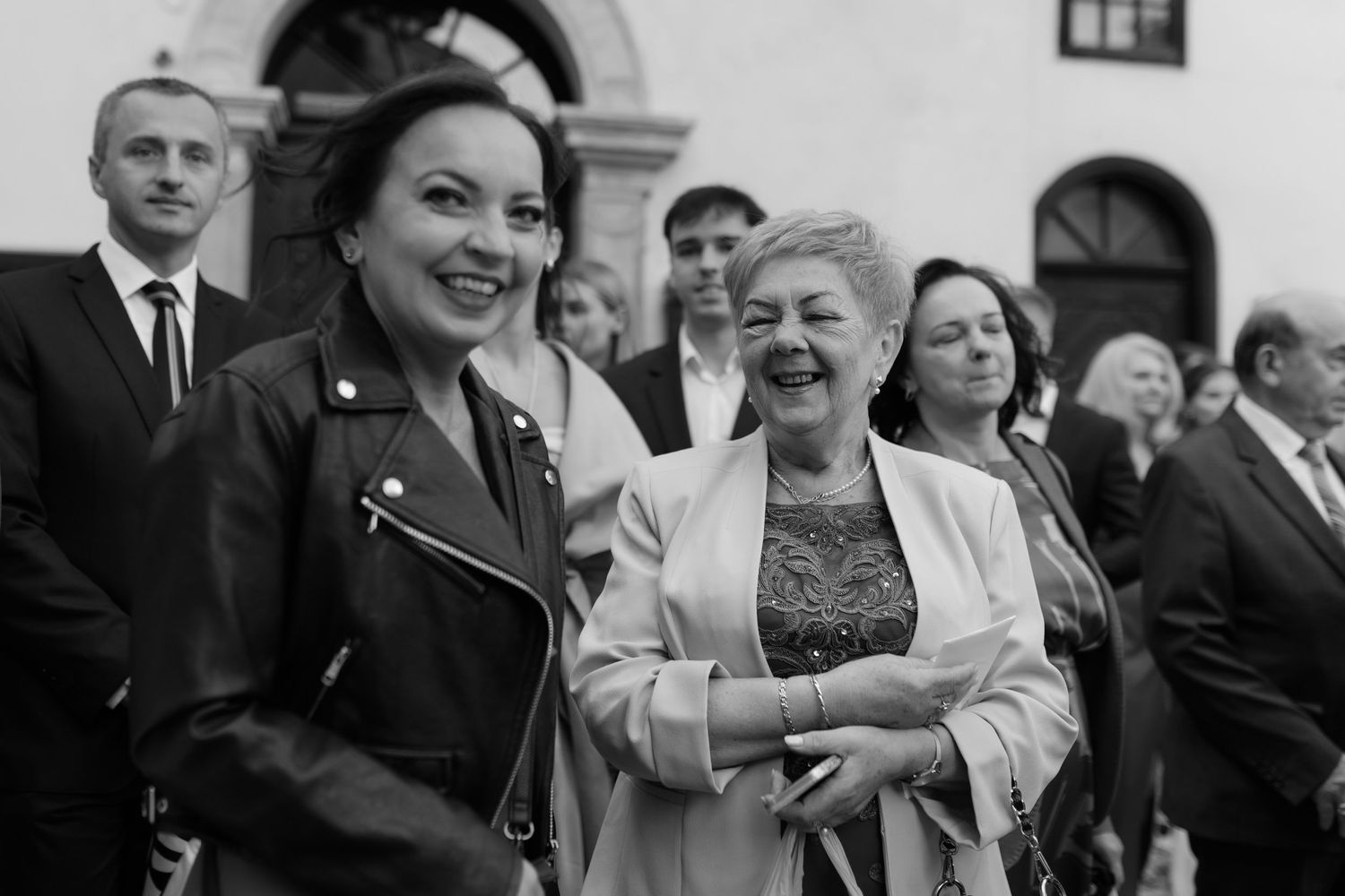 A black and white photograph shows a joyful group of people at a formal gathering, with two people in the foreground laughing.