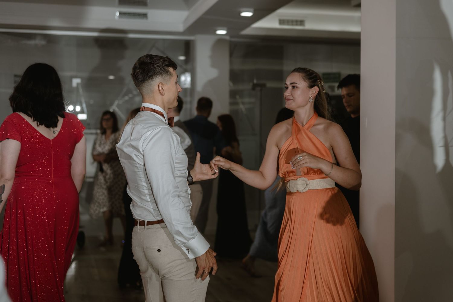 A couple wearing orange and gray attire dances together at an indoor event with other guests in the background.