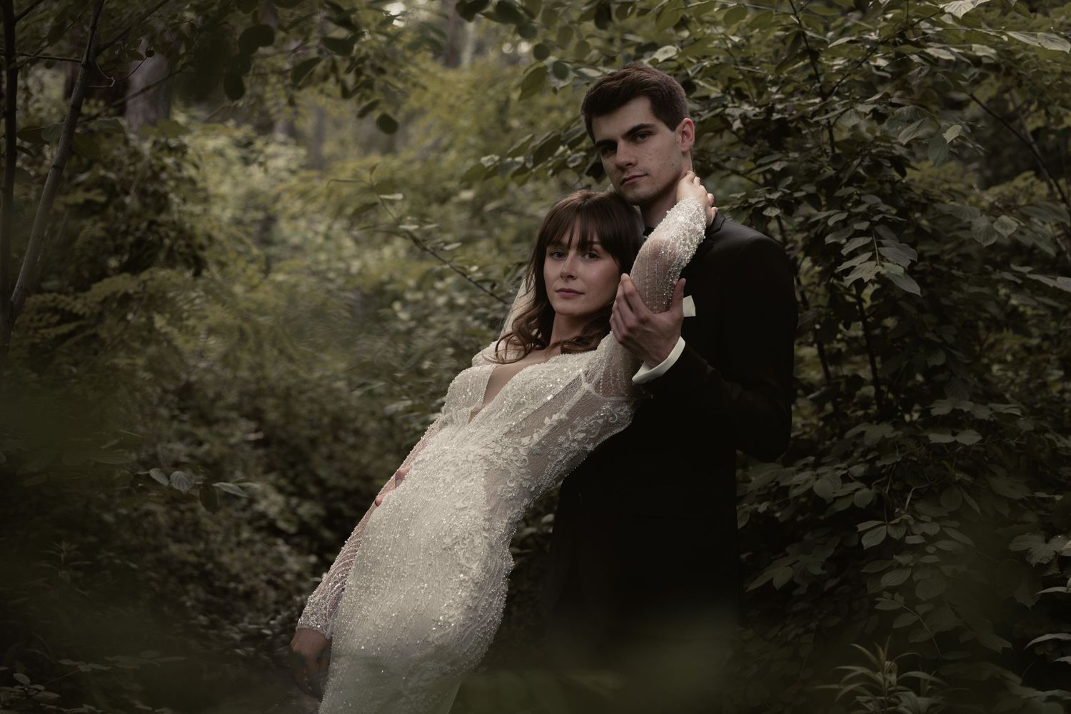 A romantic wedding couple poses together in a moody forest setting with soft lighting and lush greenery.