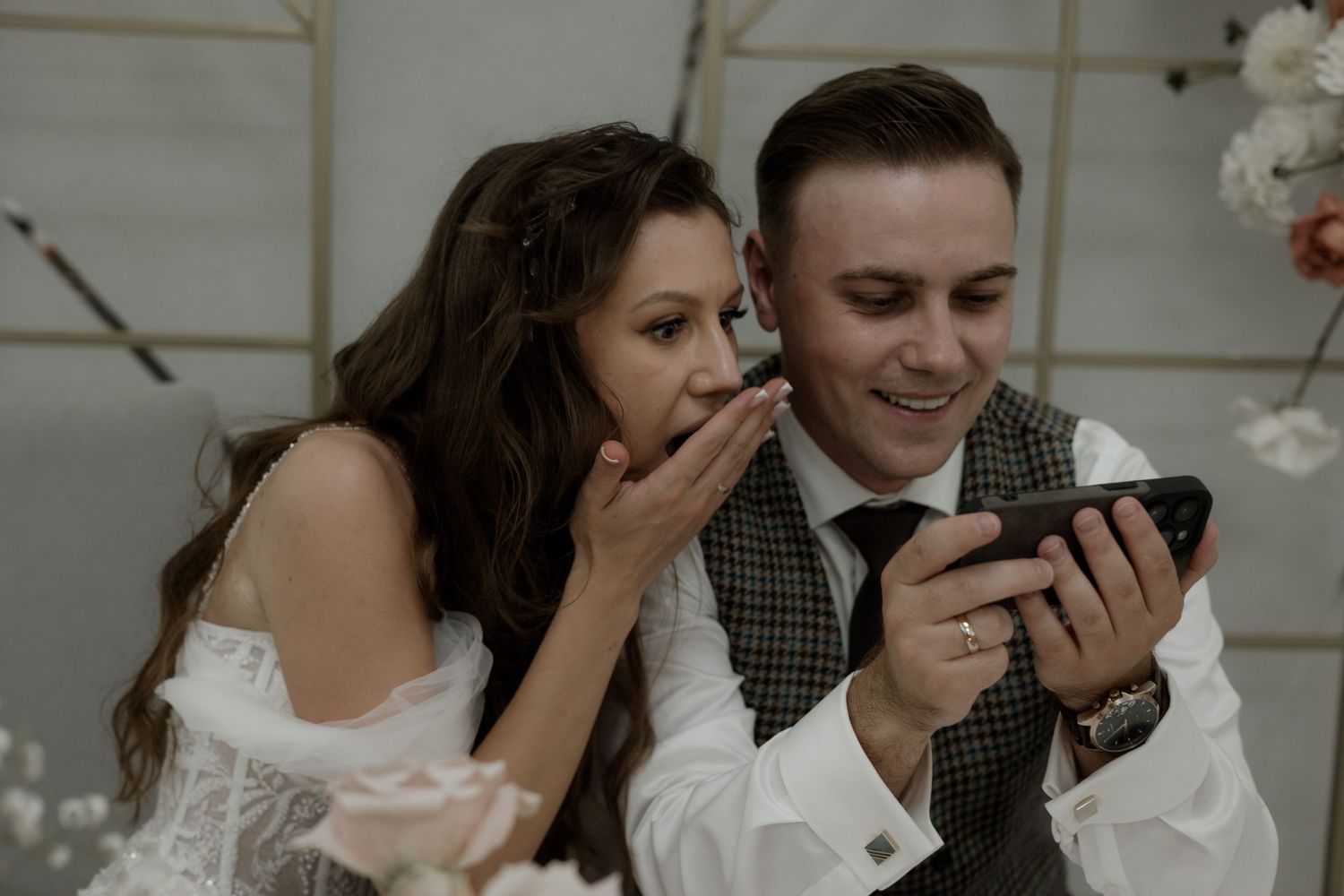 A wedding couple looks excitedly at a mobile phone during their reception.