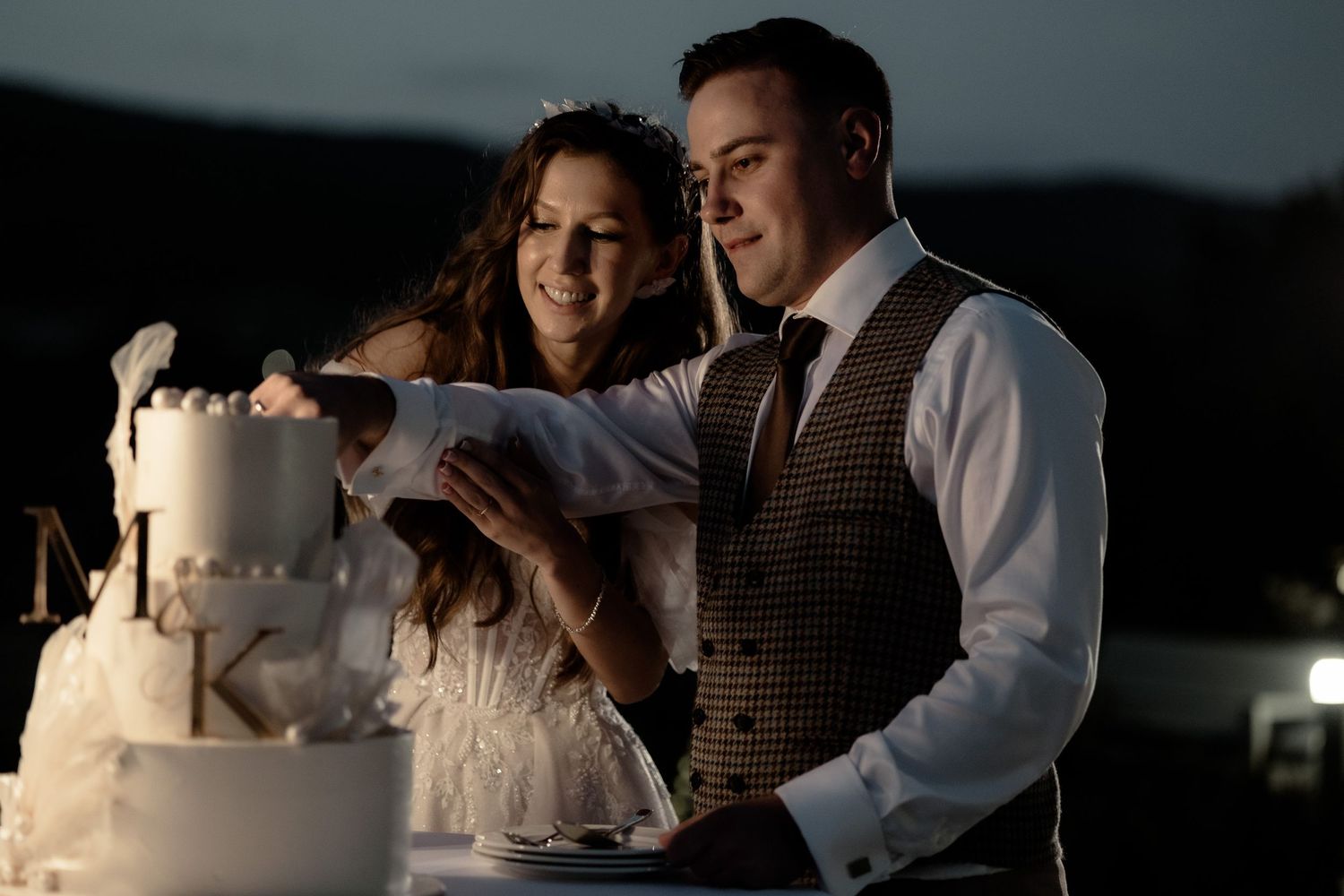 Newlywed couple cuts into their elegant white wedding cake together at an evening reception.