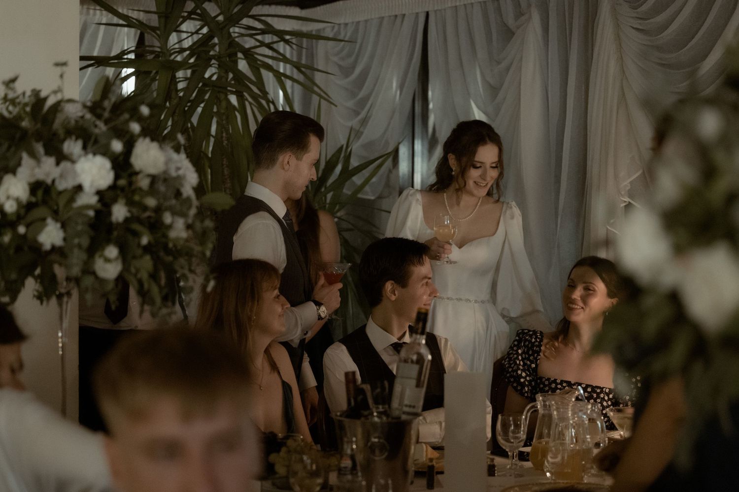 Wedding reception guests gather around an elegantly decorated table with white floral arrangements and soft lighting.