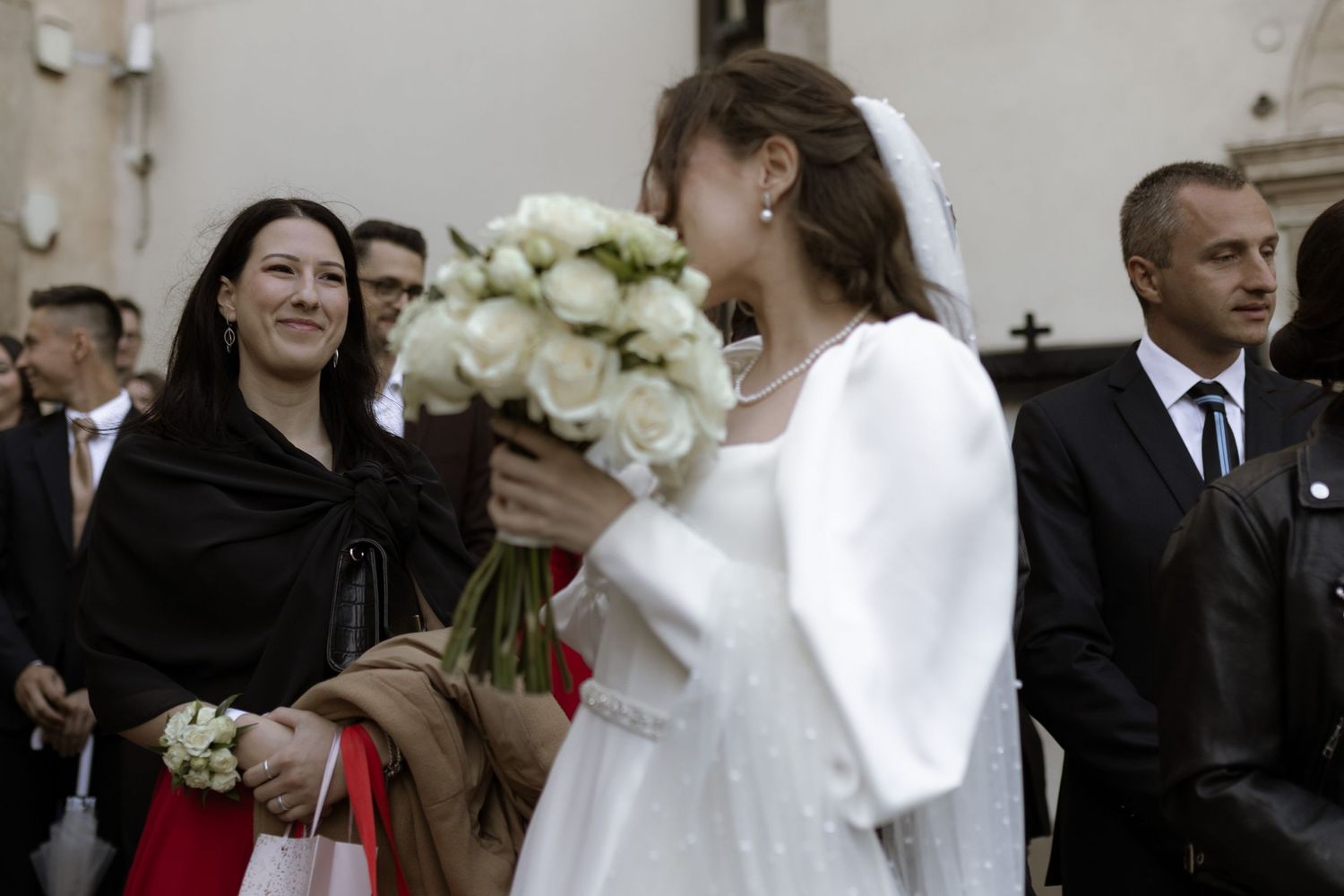 A bride in a white dress and bolero jacket holds a bouquet of white roses while guests gather outside a church.