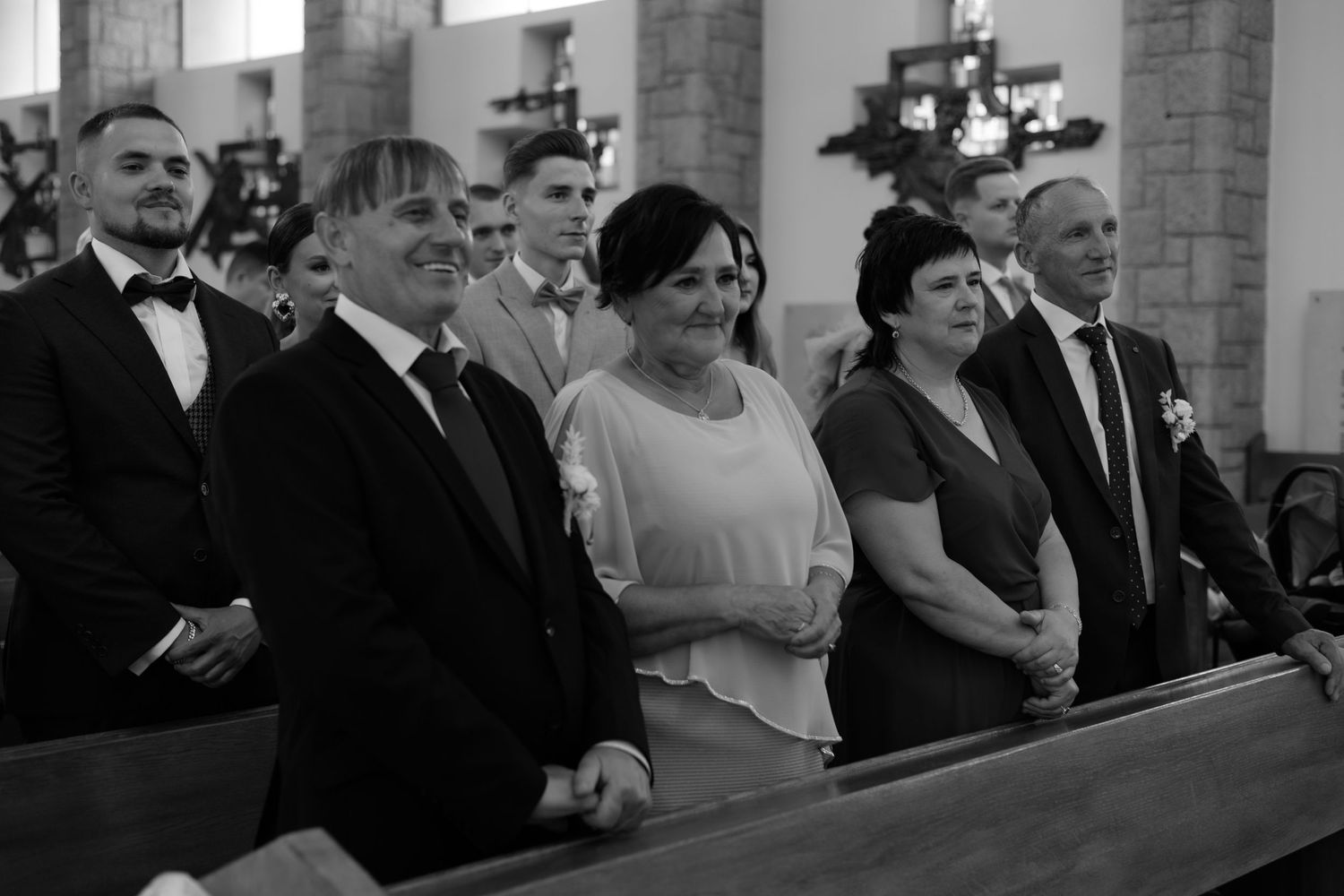 A wedding party stands solemnly in a church pew during a formal ceremony, shot in black and white.