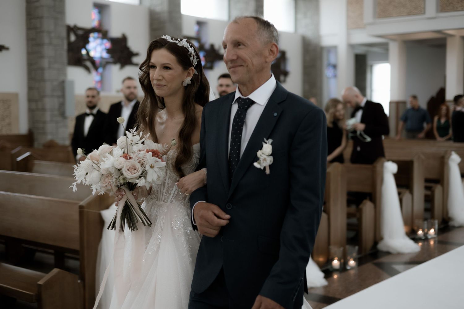 Two people stand solemnly in a church pew during a traditional wedding ceremony with white floral bouquet.