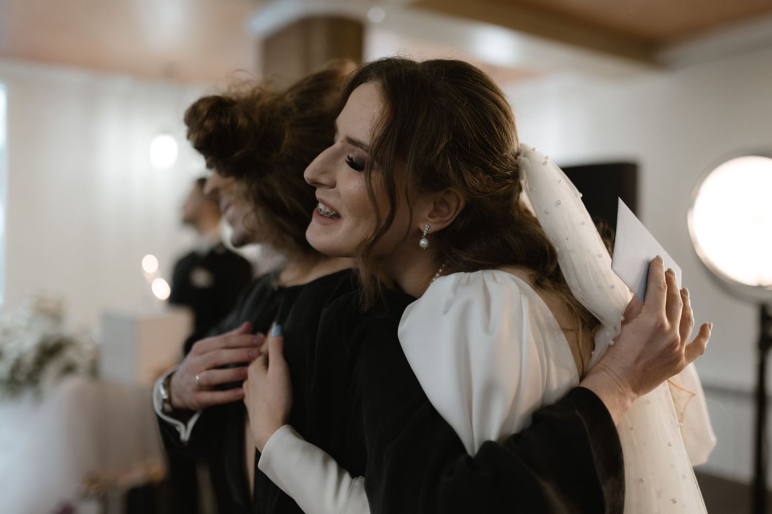 Two people share an emotional embrace at an indoor venue with soft lighting and white decor.