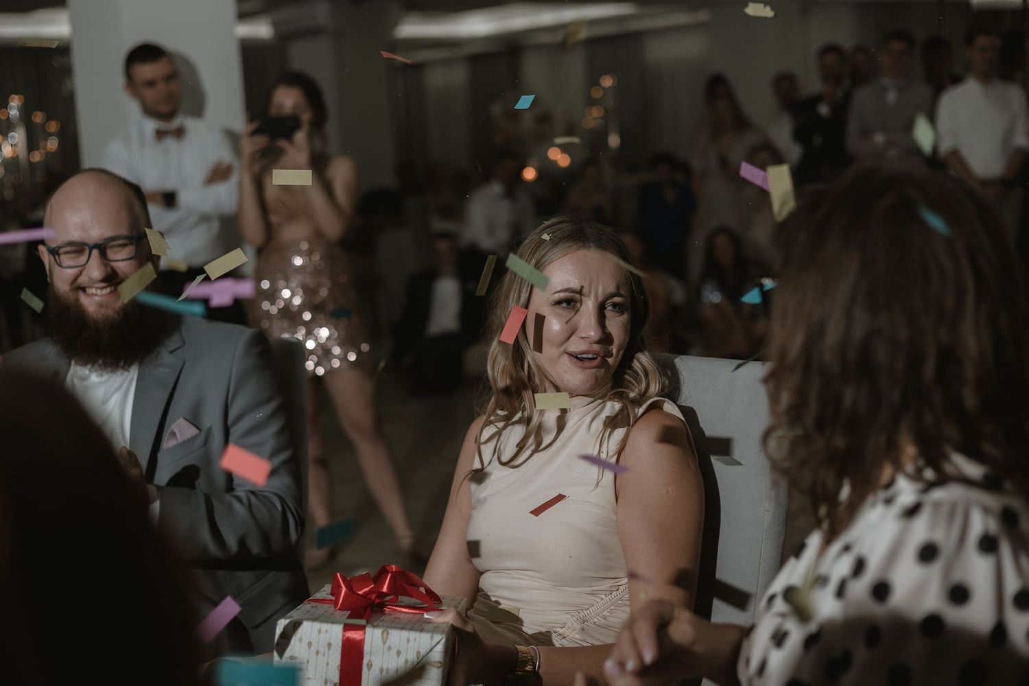 Wedding celebration scene with guests laughing at a reception table with gift boxes and festive decorations.