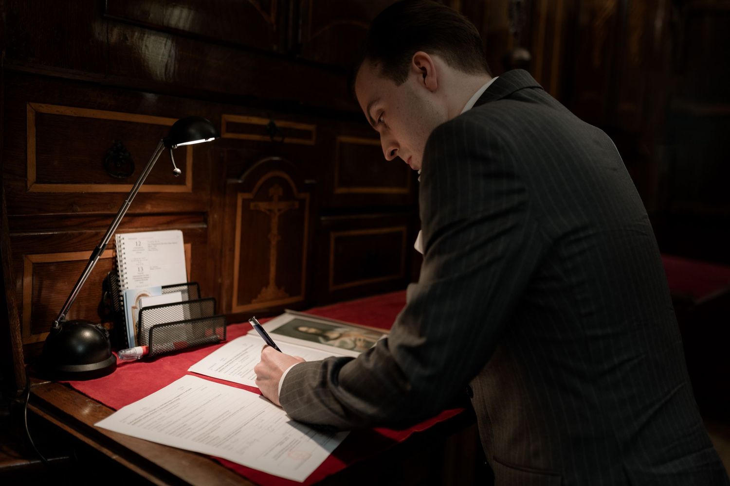 A person in a dark suit leans over a large book at a wooden desk illuminated by a small lamp.