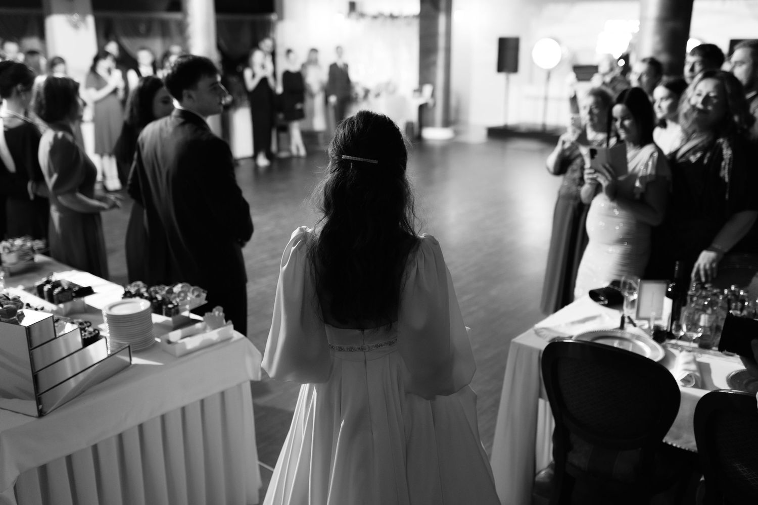 A black and white view from behind at a wedding reception showing guests gathered around tables and a buffet setup.
