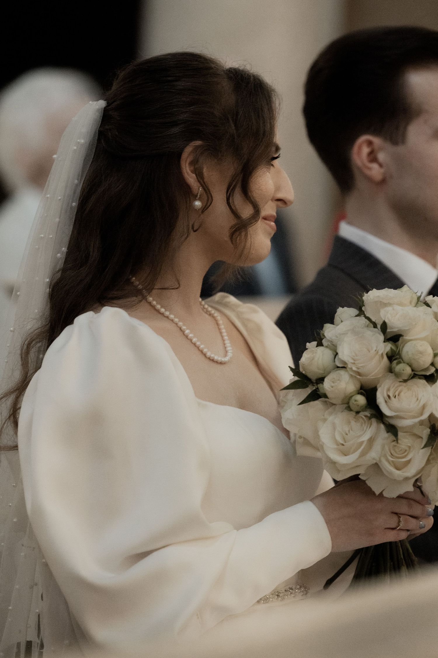 A bride in a white long-sleeved wedding dress holds a bouquet of white roses during the ceremony.