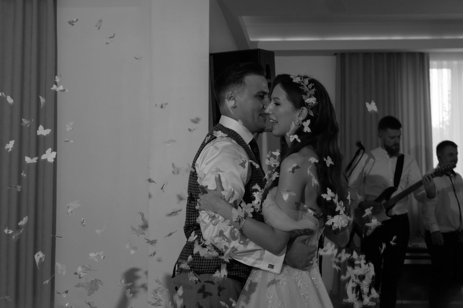 A romantic black and white wedding dance moment with floating petals in the air while musicians perform in the background.