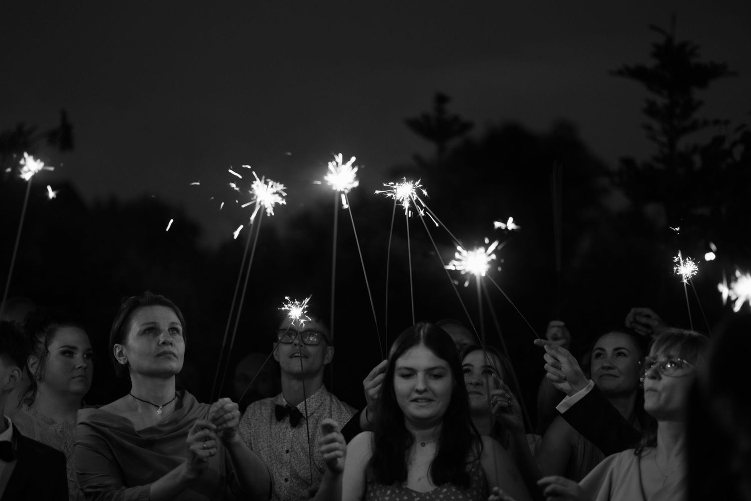 A black and white nighttime photo shows a group holding bright sparklers against a dark sky.