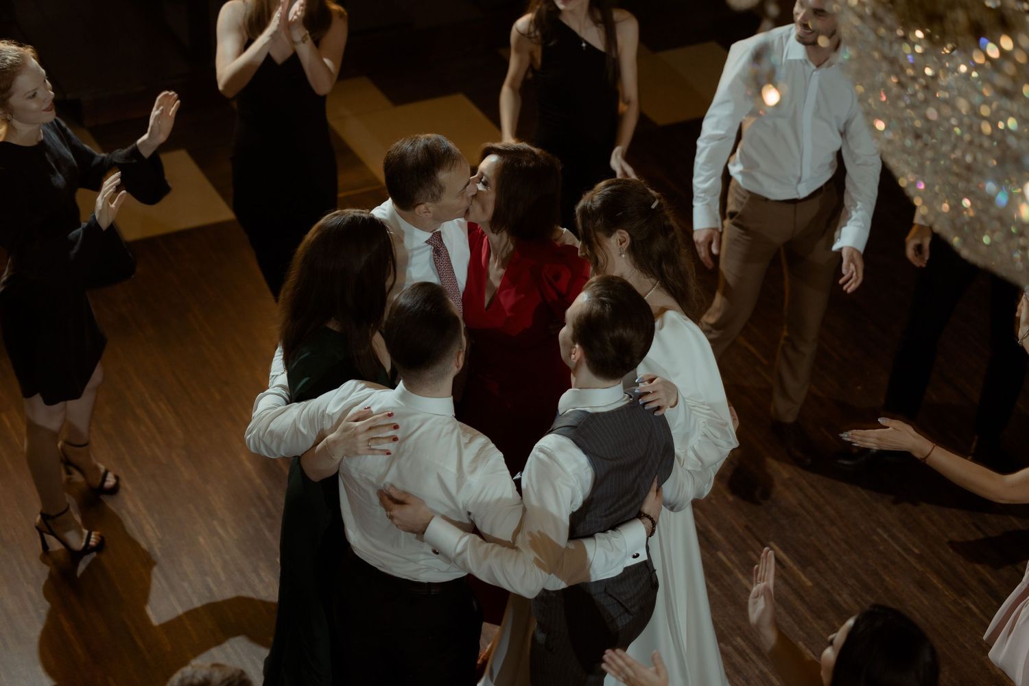 People gathered in a circle on a wooden dance floor during a celebration with twinkling lights in the background.