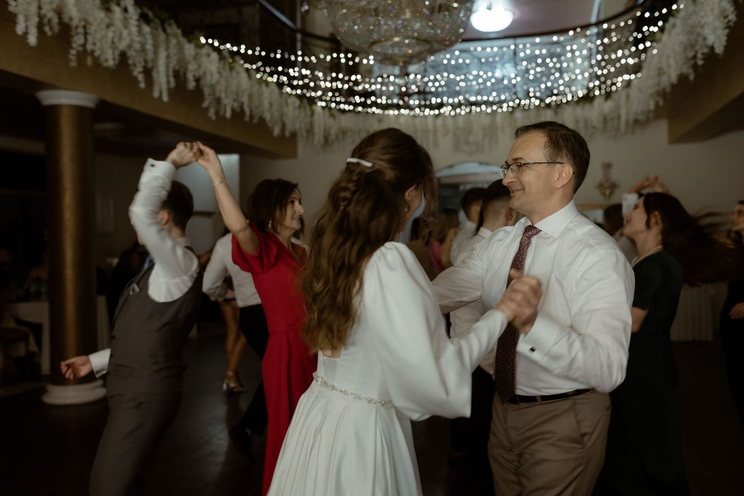 People dancing at a reception under twinkling string lights and white ceiling drapes in an indoor venue.
