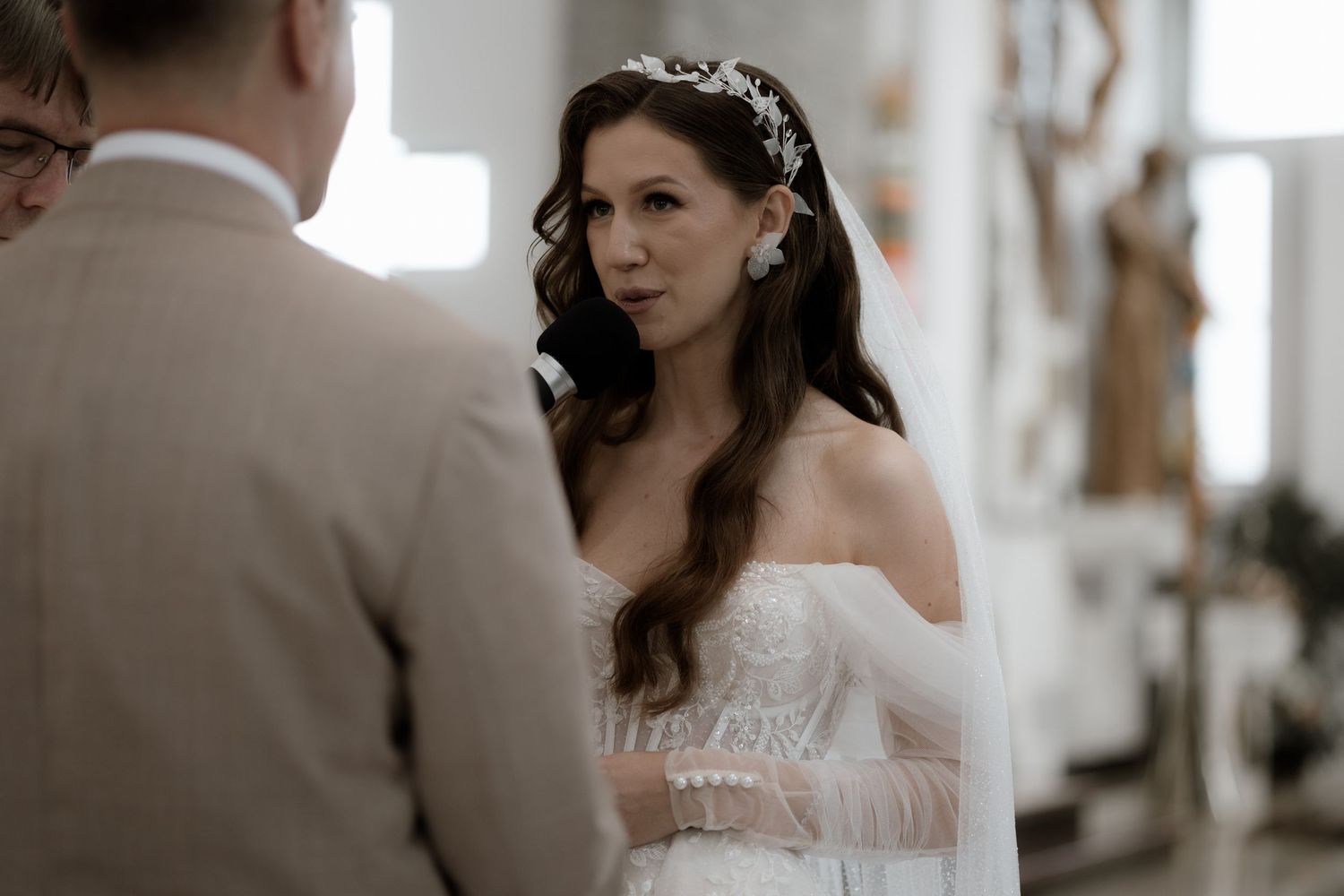 A bride in a white off-shoulder wedding gown with long wavy hair and delicate crown looks lovingly during ceremony.