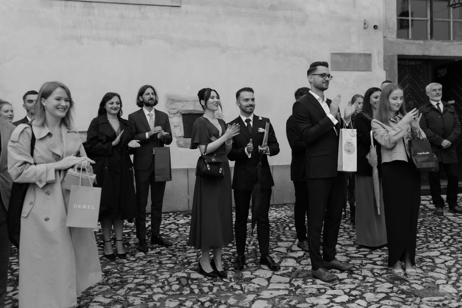 A group of well-dressed people stand together on a cobblestone surface, appearing to be at a formal outdoor gathering.