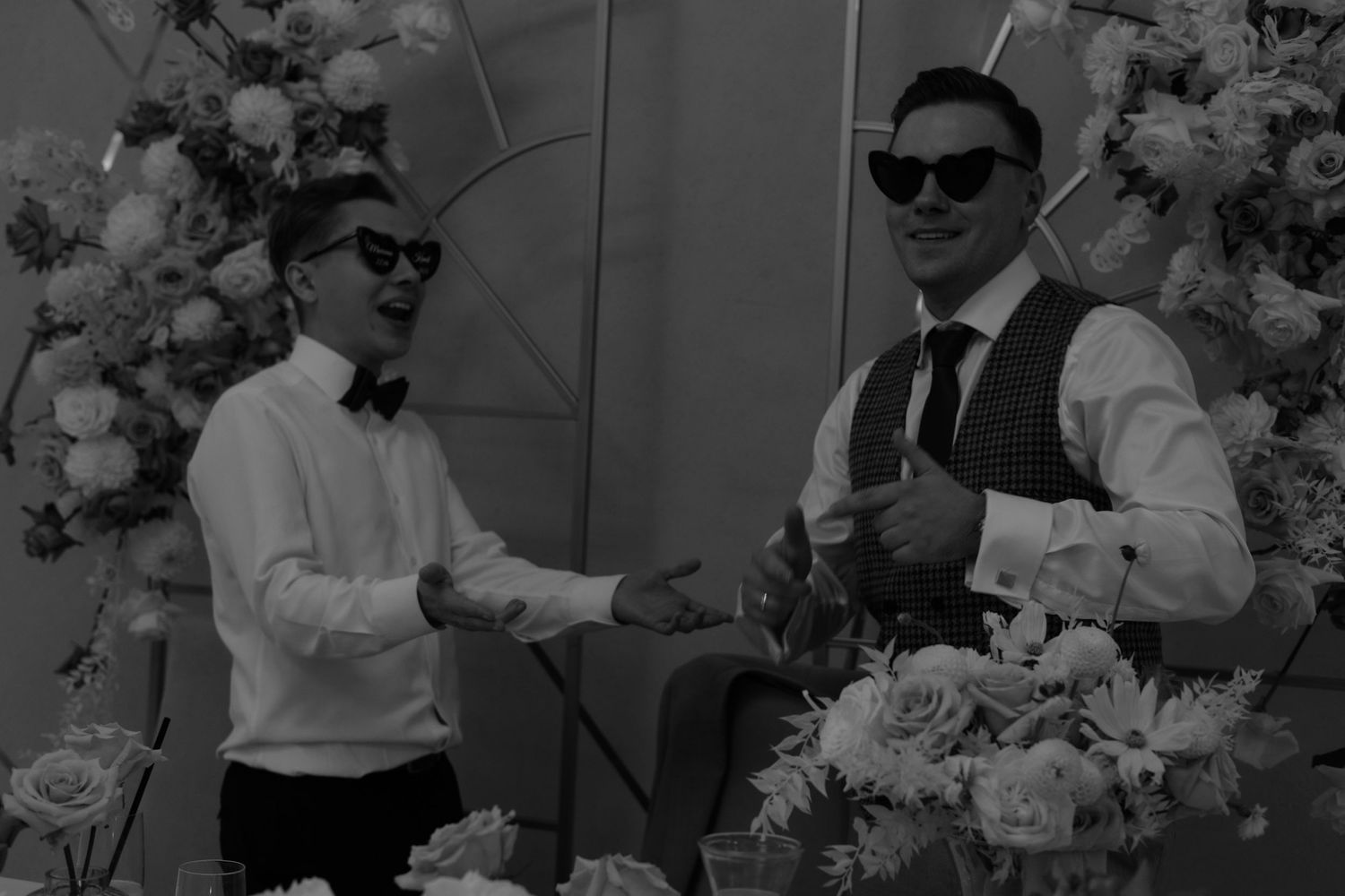A black and white image of two people in formal attire standing amidst floral decorations.