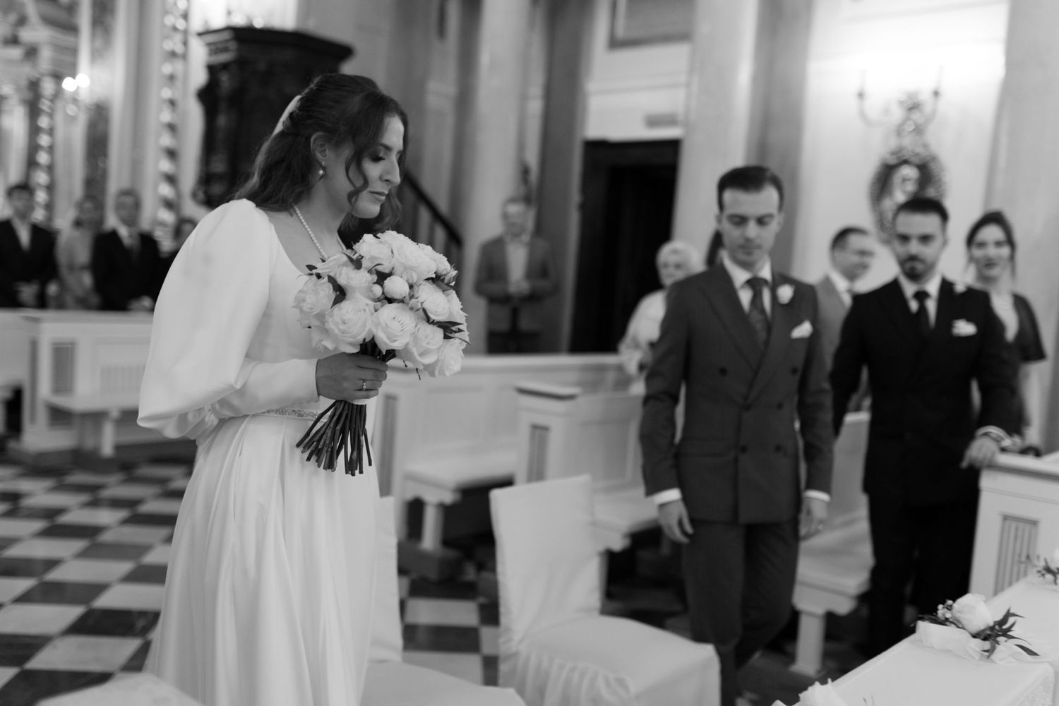 A black and white wedding photo showing a bride holding a bouquet of white flowers in an elegant room with checkered floors.
