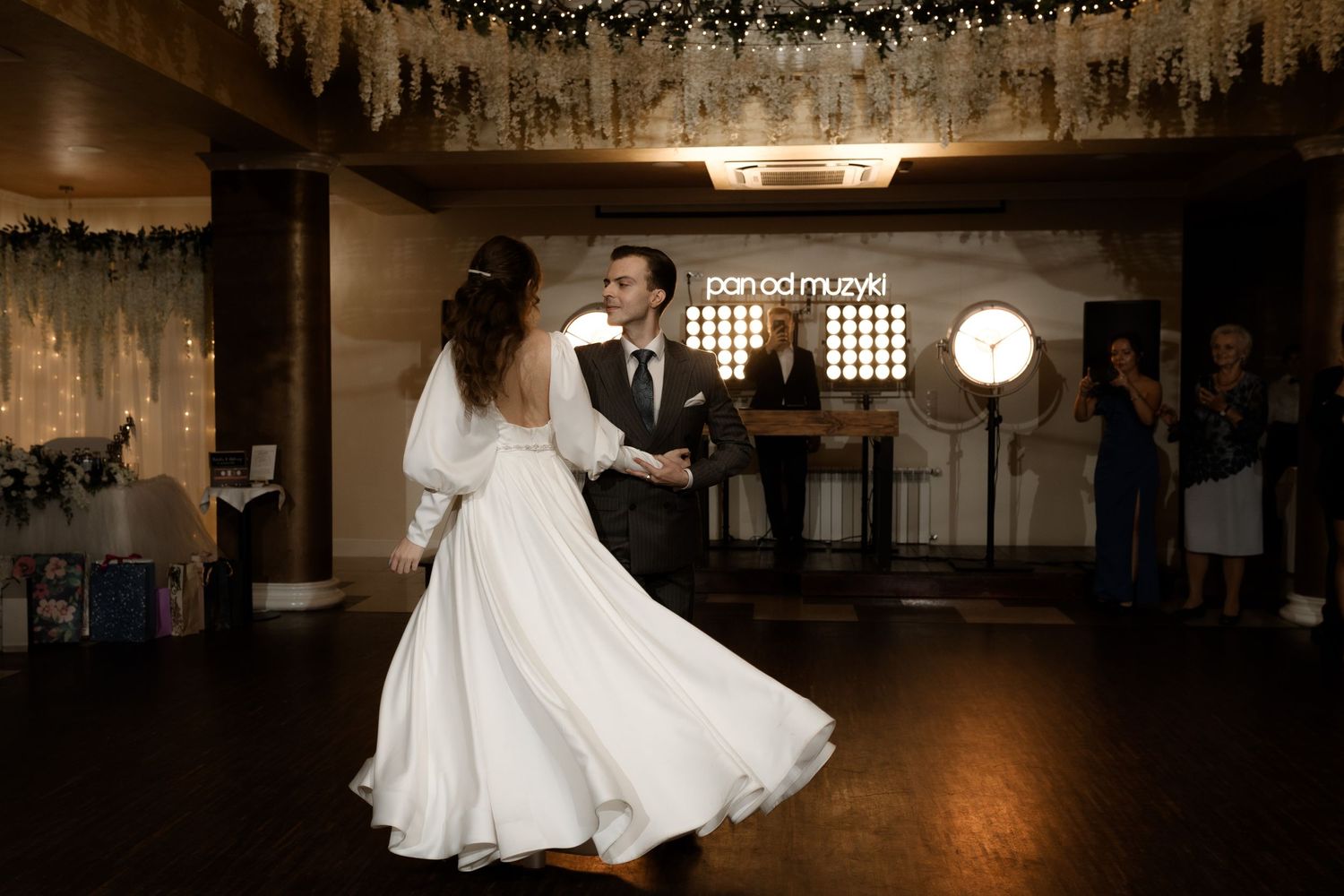 A couple shares their first dance under hanging floral decor at a romantic wedding reception venue with rustic lighting.