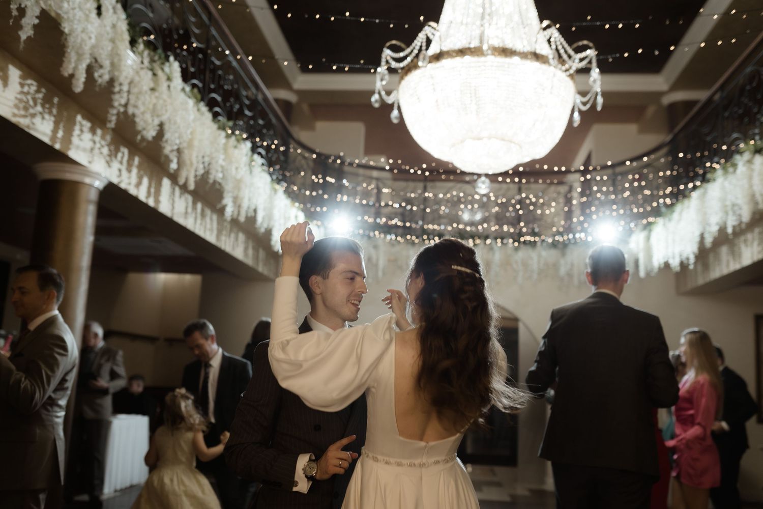 A couple dances together under a sparkling chandelier and twinkling lights at an elegant wedding reception.
