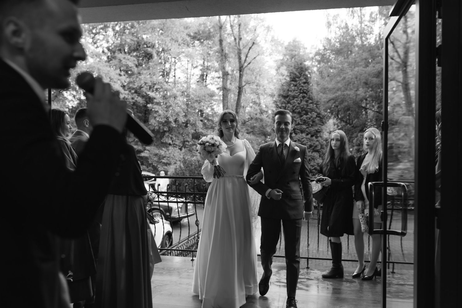 A black and white outdoor wedding ceremony takes place on a porch surrounded by trees.