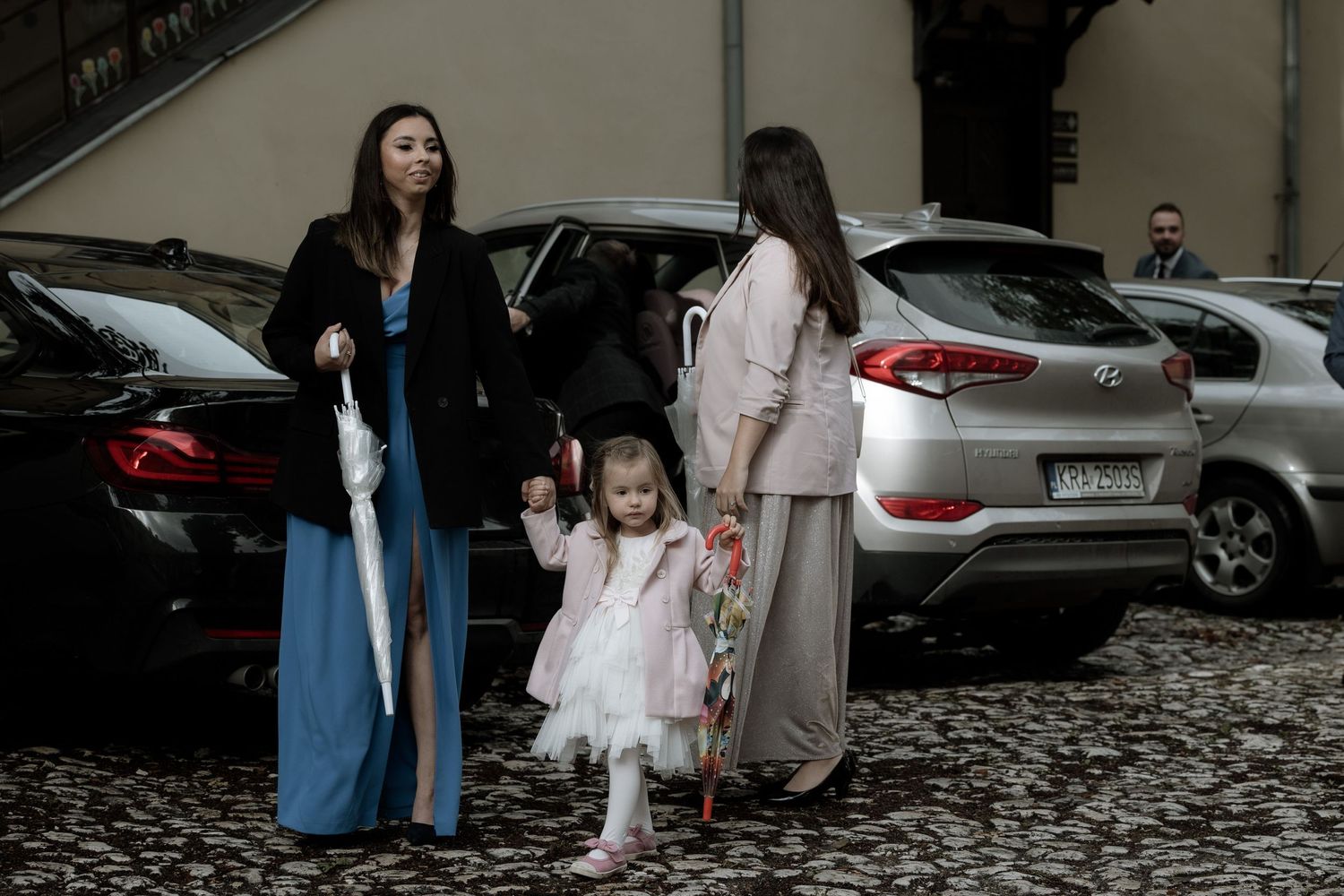 Group gathered near parked cars in a cobblestone area, dressed in formal attire including a blue dress and pink dress.