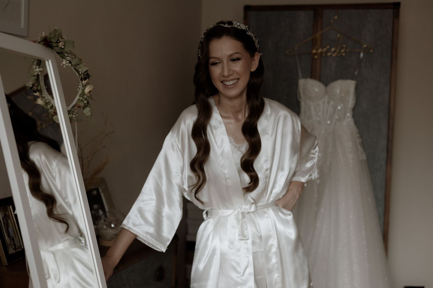A woman in a white silk robe prepares on her wedding day with a wedding dress visible hanging in the background.
