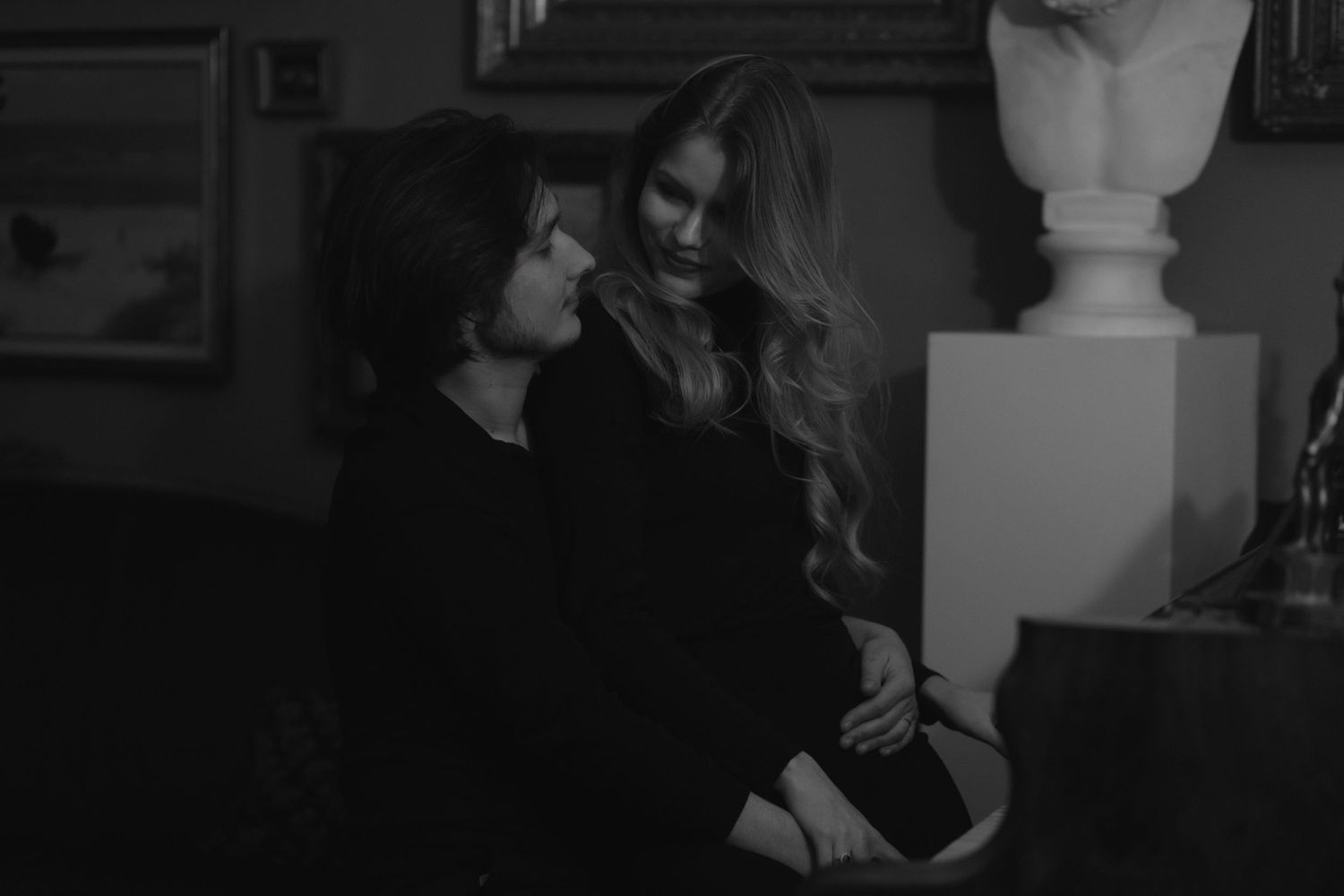 A black and white photograph shows two people in dark clothing talking intimately next to a decorative bust.
