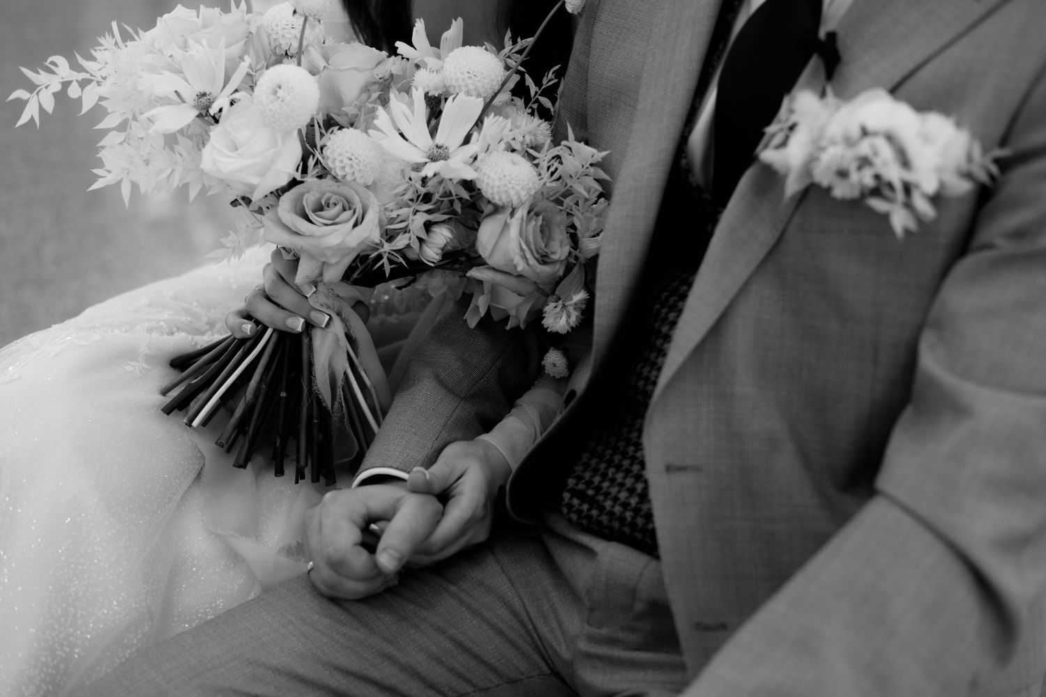 Black and white close-up of wedding rings and bouquet with roses and daisies on a bridal gown.