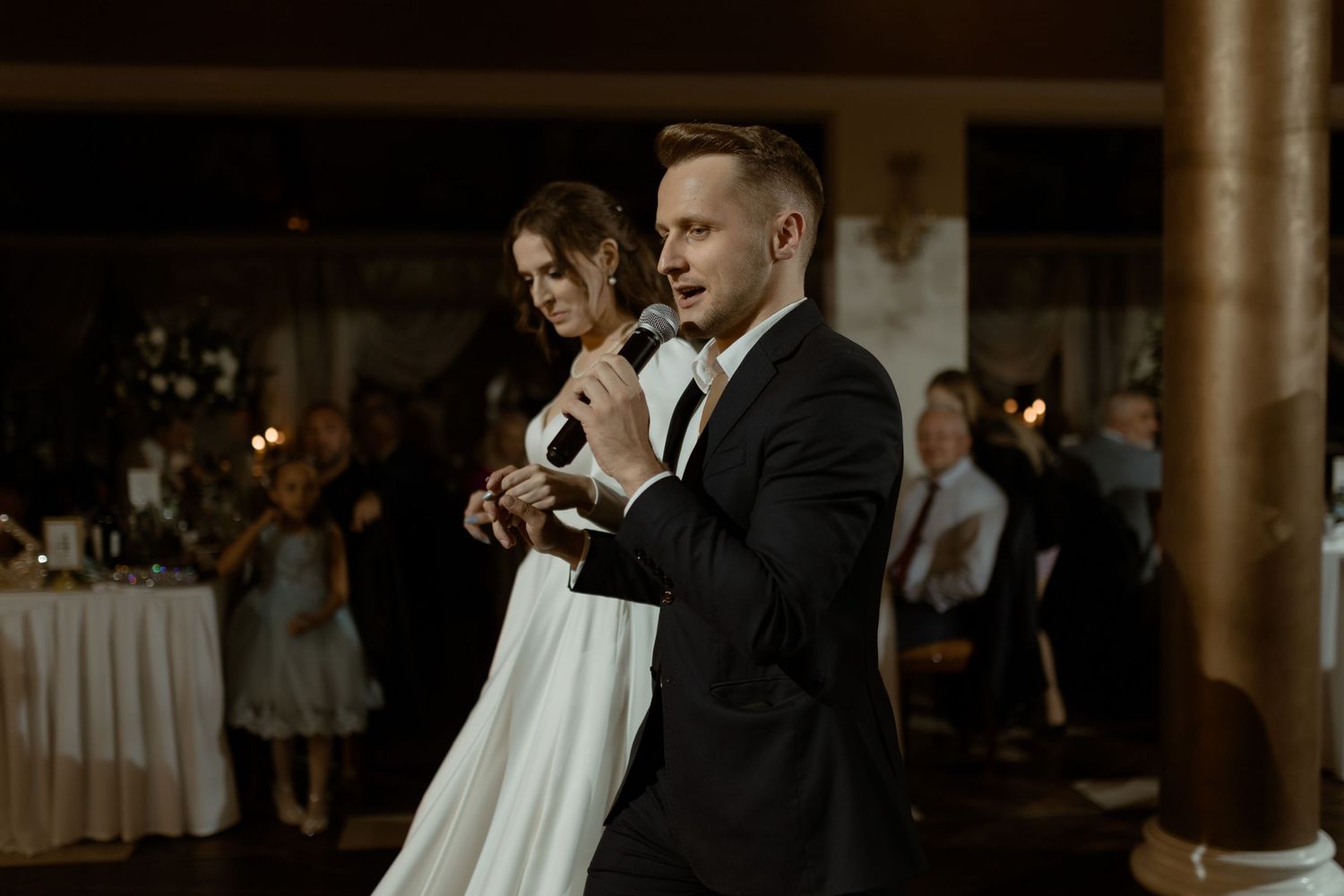 Two formally dressed guests speak into microphones at an elegant indoor wedding reception venue.